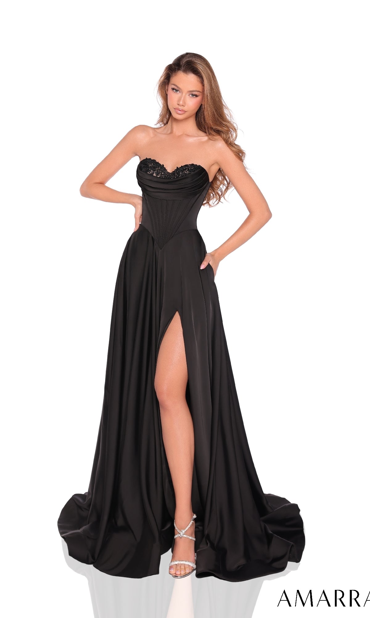 A woman models the Long Prom Dress: Amarra 89023, a strapless black gown with a corset bodice, sweetheart neckline, high slit, and silver heels, posed against a white background.